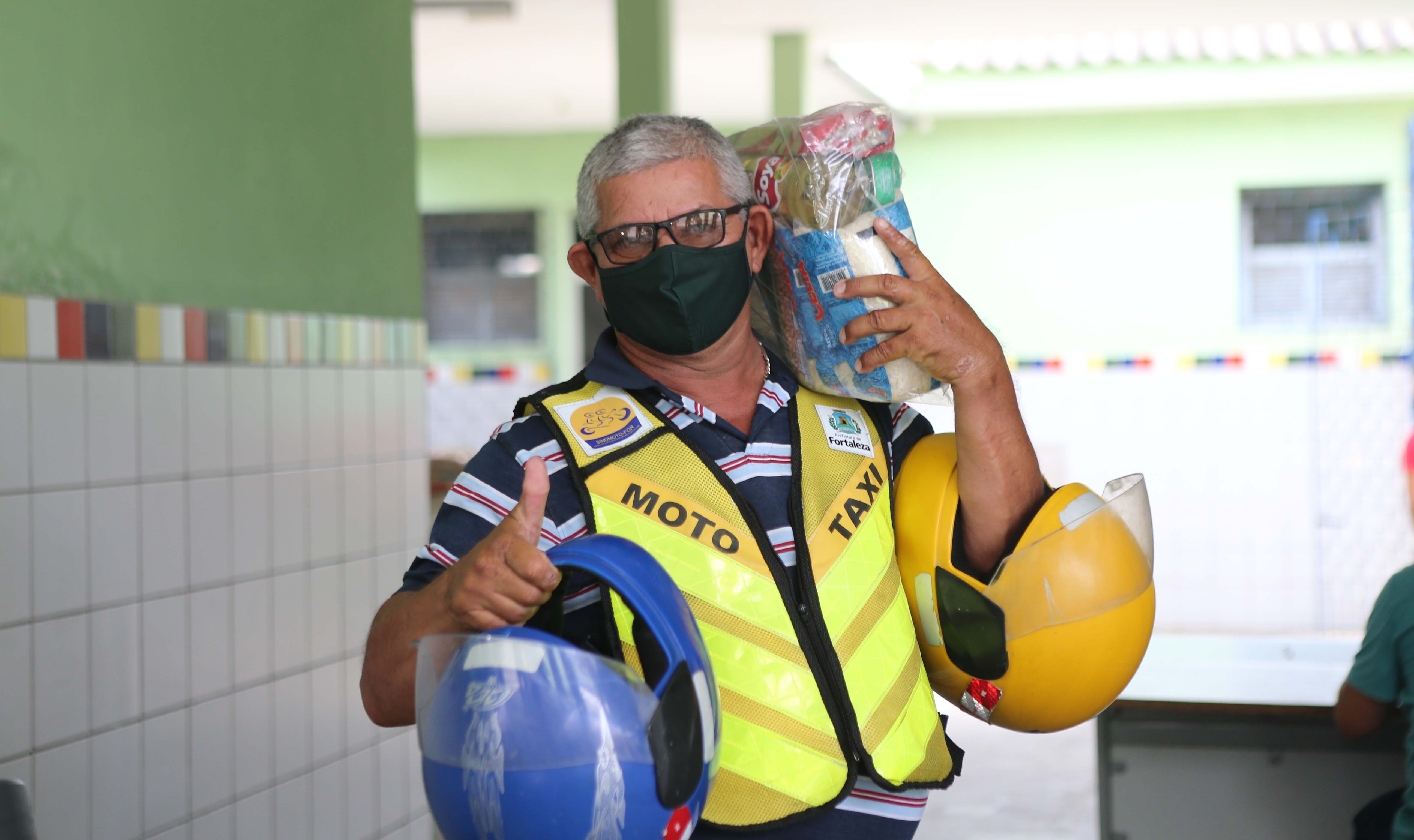 homem usando colete de mototaxista faz sinal de positivo para a câmera enquanto segura uma cesta básica